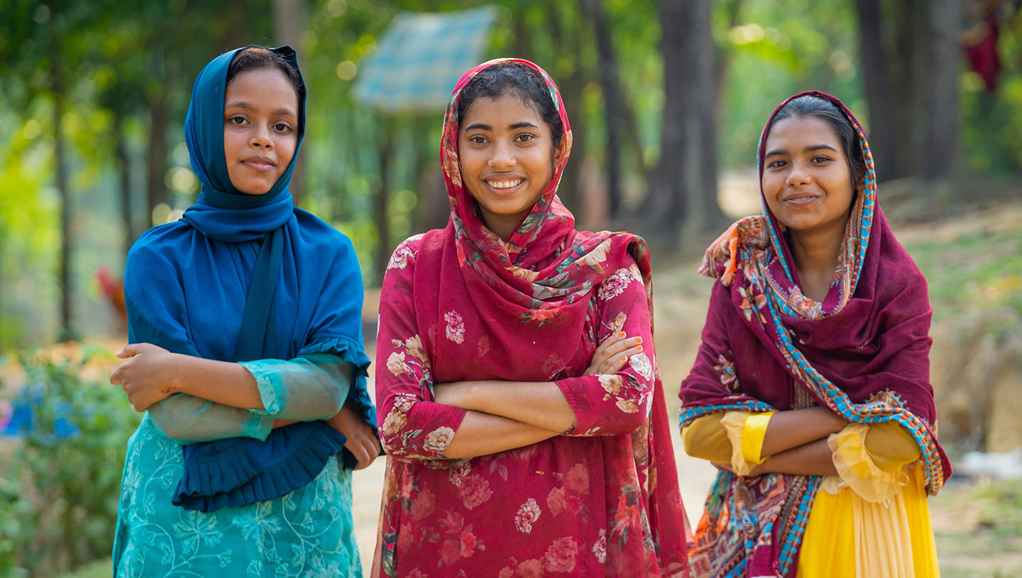 Three young girls stand outdoors with arms crossed, smiling warmly. They're wearing colorful shawls and dresses. The background is a lush, sunlit park.